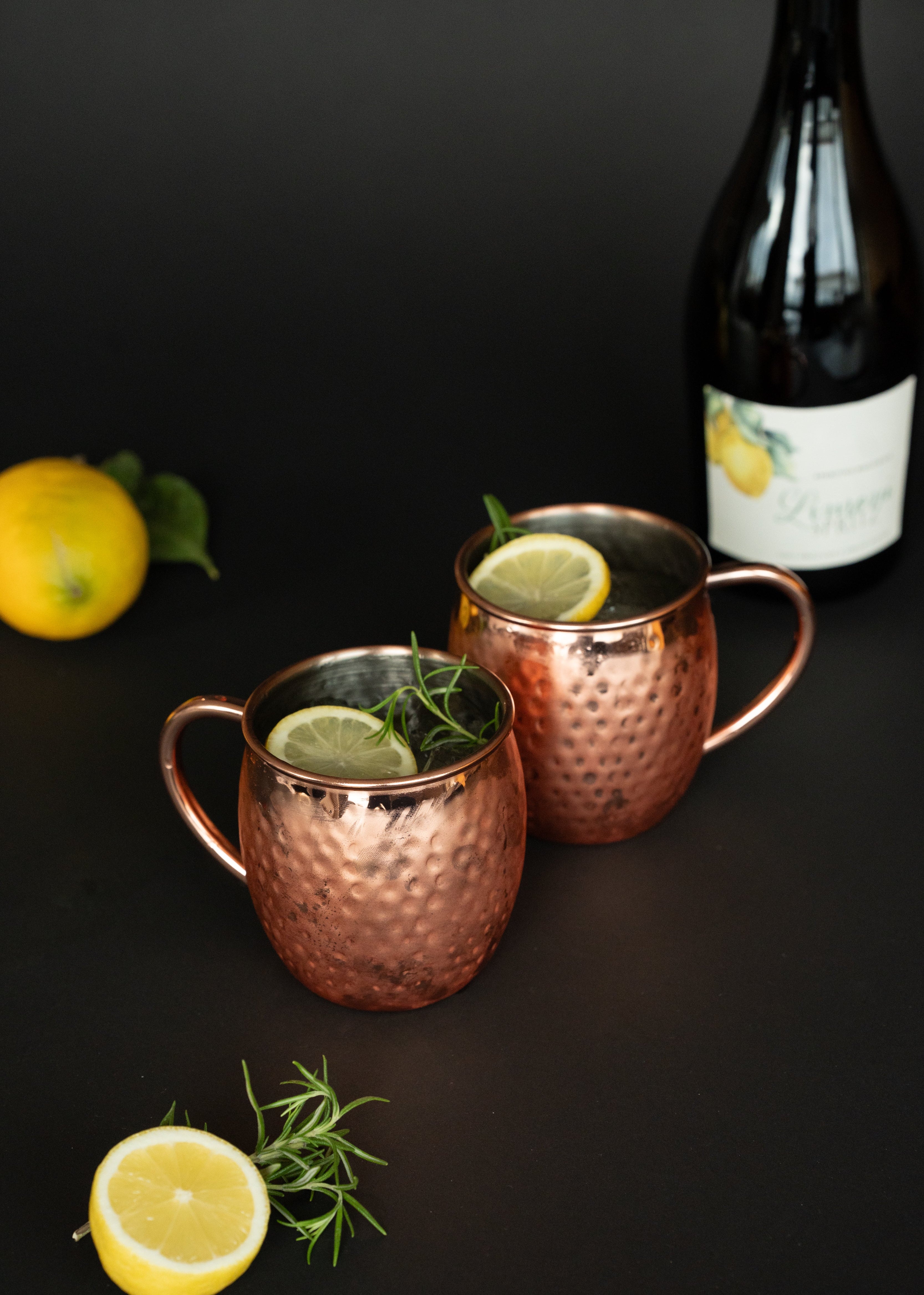 2 Kupferbecher gefüllt mit Limoni Spritz, einer Zitronenscheibe und Rosmarin. Flasche steht im Hintergrund