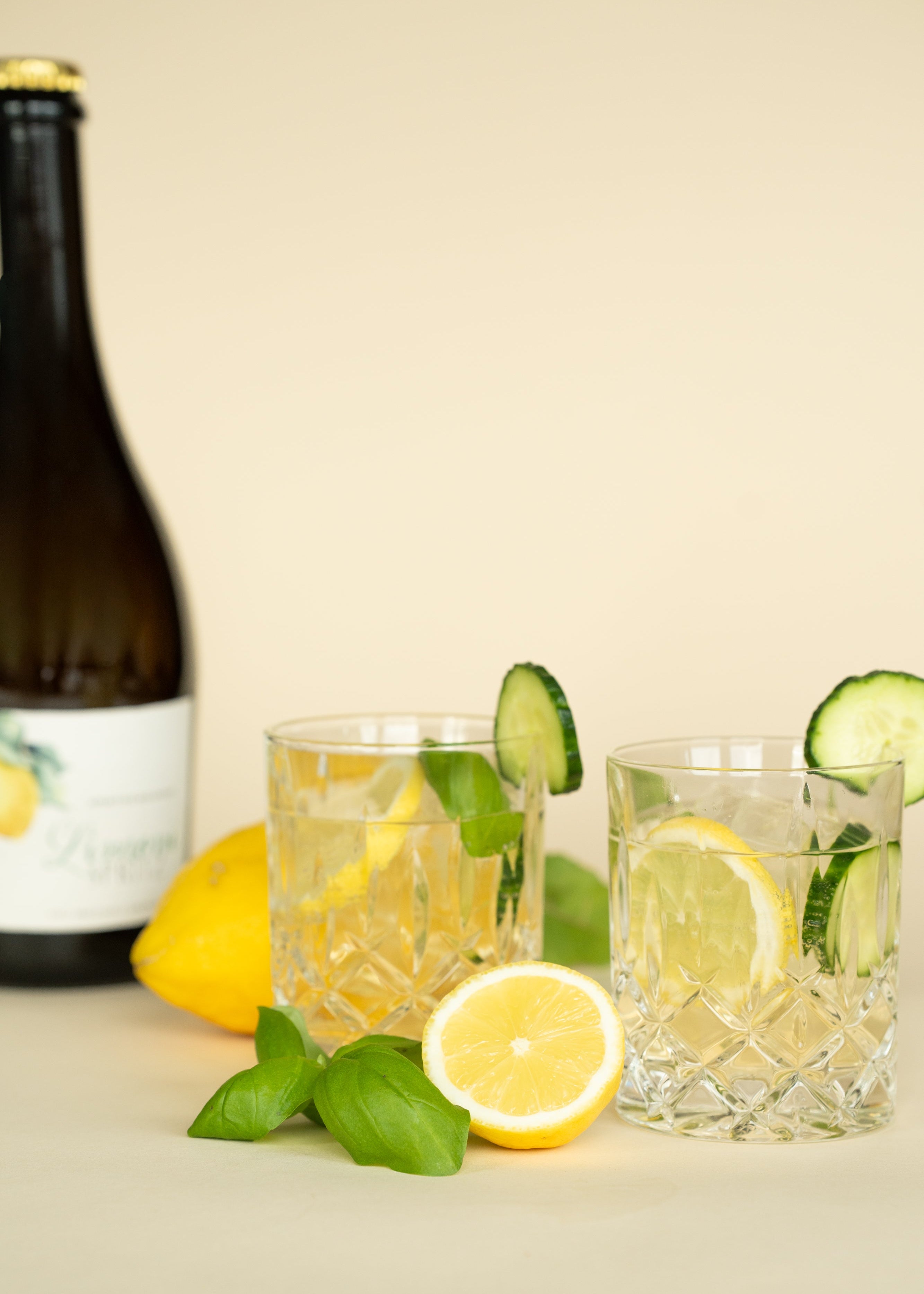 Zwei Gläser Limoni Spritz mit Zitrone, Gurke und Basilikum und einer Flasche Limoni Spritz im Hintergrund