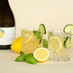 Zwei Gläser Limoni Spritz mit Zitrone, Gurke und Basilikum und einer Flasche Limoni Spritz im Hintergrund