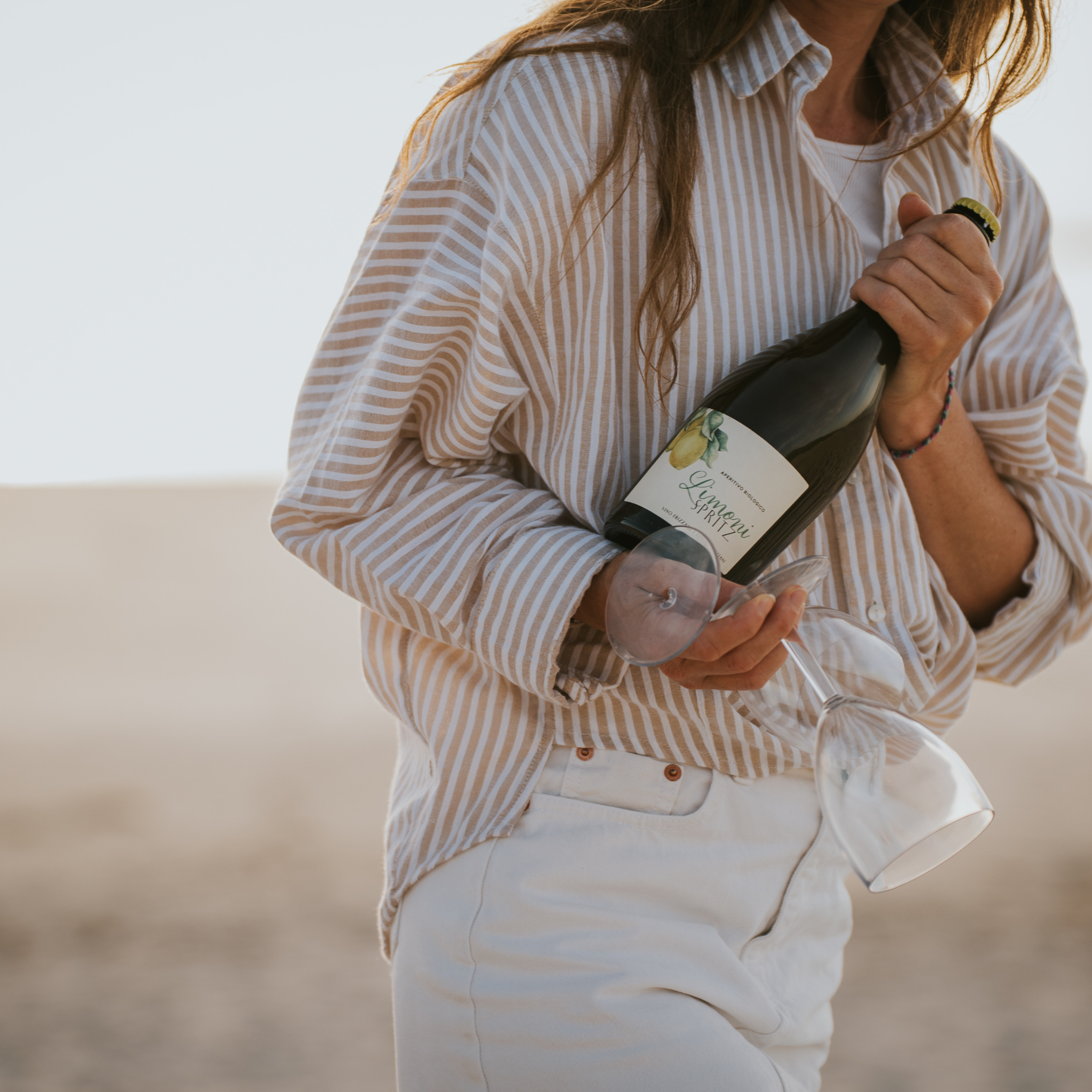 Frau am Strand mit einer gestreiften Bluse die eine Flasche Limoni Spritz und 2 Weingläser in den Händen hält