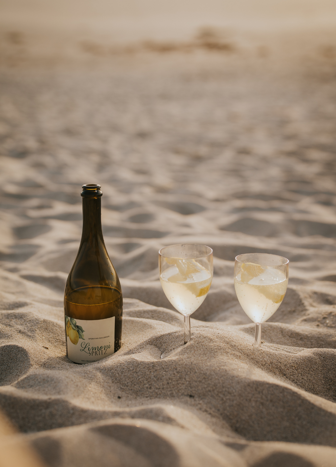 Geöffnete Limoni Spritzflasche und 2 gefüllte Weingläser die am Strand im Sand stehen.