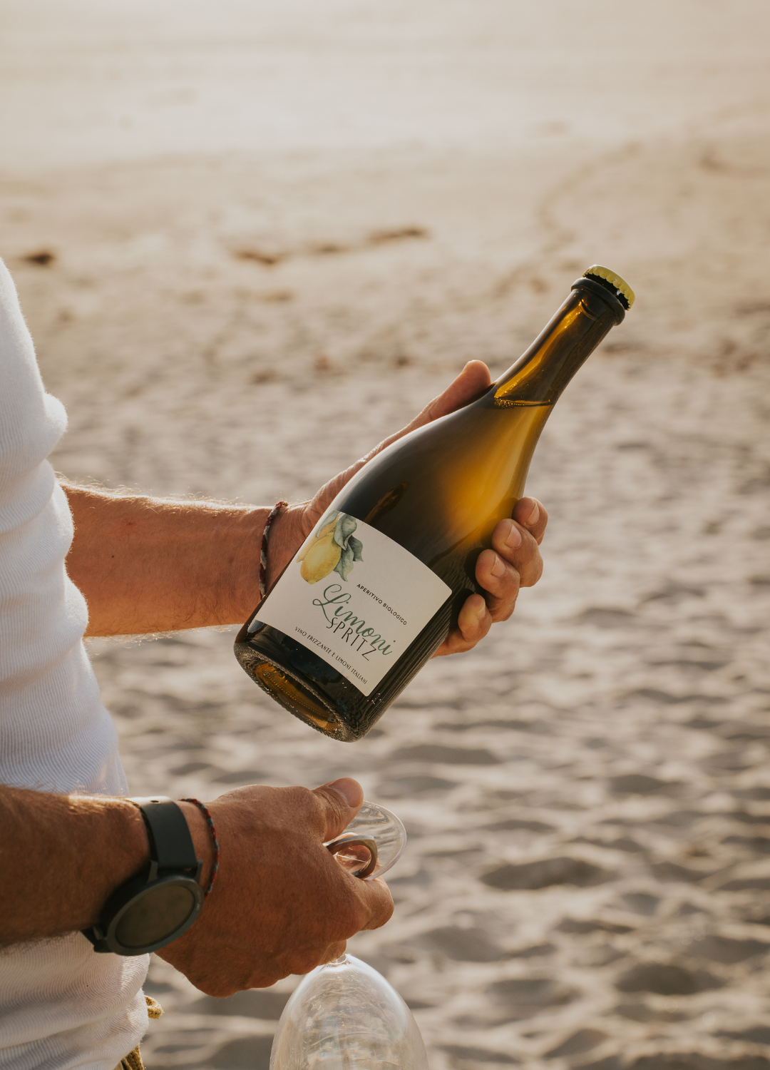 2 Männerhände am Strand die in der linken Hand eine Flasche Limoni Spritz hält und in der rechten ein Weinglas.